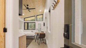 A narrow hallway leading to a bedroom with a bed, a small table, and chairs. There are large windows and a ceiling fan. - Guanella Studio Cabin | FirePit | Creek | Lostfork (Grant)