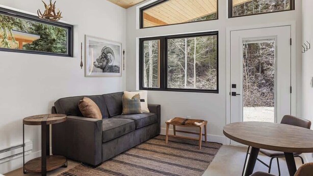 A modern living room with a dark grey sofa, a round wooden coffee table, a small wooden side table, and a round dining table with chairs. Large windows provide views of trees and a snowy landscape. - Exclusive Basecamp Buyout | 14 Cabins | Lostfork (Grant)
