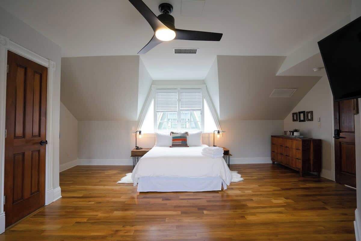A bedroom with a large bed, a ceiling fan, a window with blinds, and a wooden dresser.