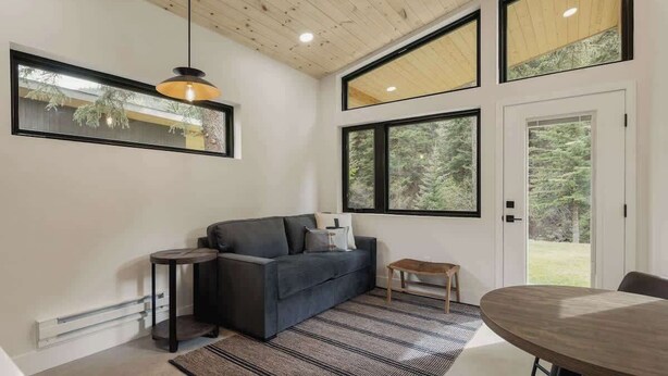 A modern living room with a dark grey sofa, a small round table, a hanging pendant light, and large windows offering a view of greenery. - Guanella Cabin Escape | Hot Tub | Sauna | Lostfork (Grant)