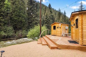 A wooden cabin with a deck overlooking a river, surrounded by dense forest. - Guanella Cabin Escape | Hot Tub | Sauna | Lostfork (Grant)