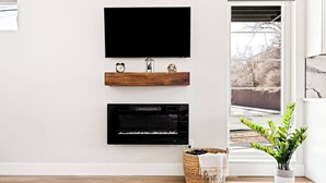 A modern living room with a flat-screen TV mounted above a wooden shelf, a fireplace below, and a view of a snowy landscape through a large window. - Golden Sanctuary | Luxe Apt | 1 Block From Main St (Golden)