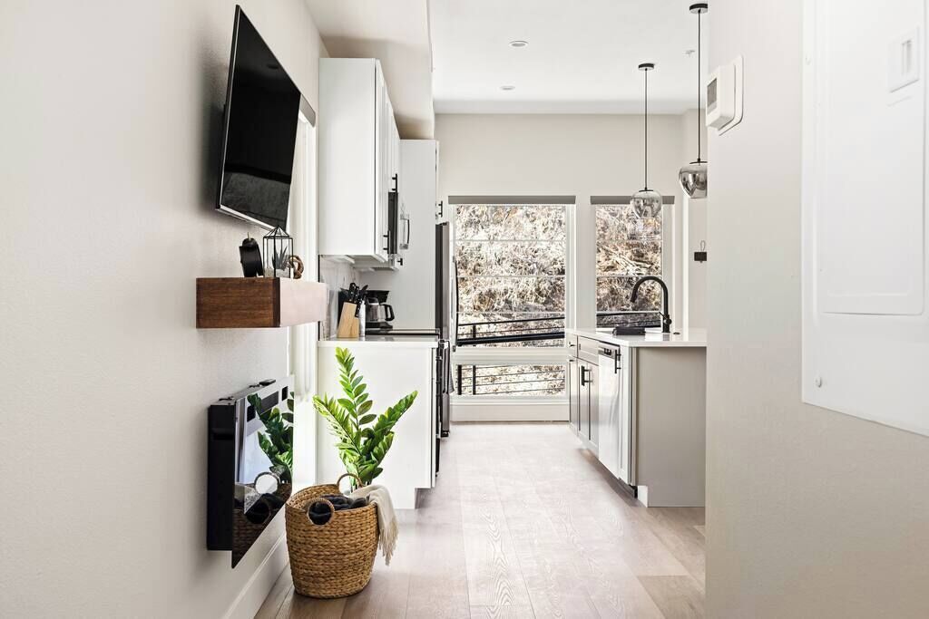 A modern kitchen with white cabinets, a TV mounted on the wall, and a large window offering a view of trees.