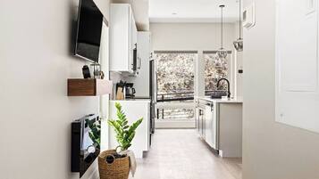 A modern kitchen with white cabinets, a TV mounted on the wall, and a large window offering a view of trees.