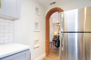 Uma cozinha com geladeira de aço inoxidável, armários brancos, um arco de tijolos e uma área de jantar ao fundo.