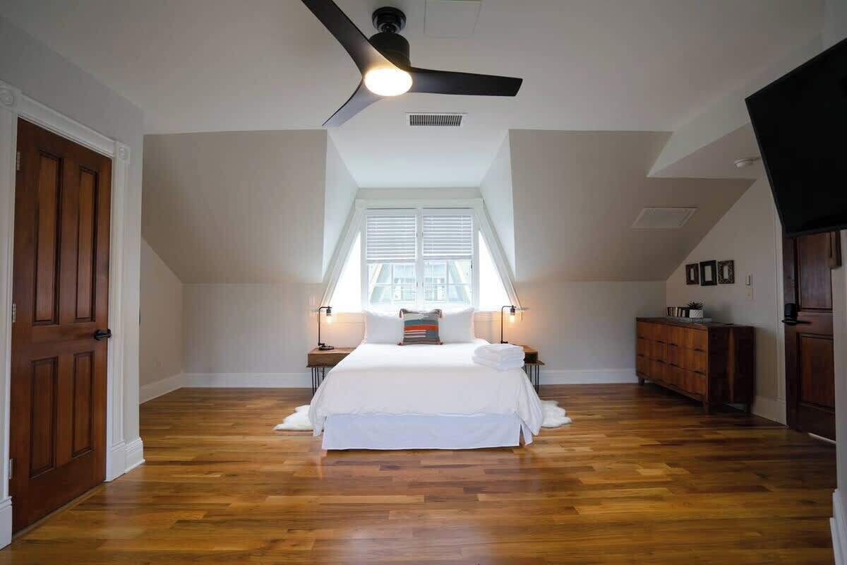 A bedroom with a wooden floor, a ceiling fan, a bed with white bedding, a window with blinds, and a nightstand with a lamp.