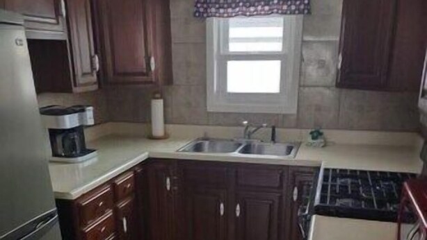 A compact kitchen with dark wooden cabinets, a stainless steel refrigerator, a coffee maker, a sink, and a patterned window valance.