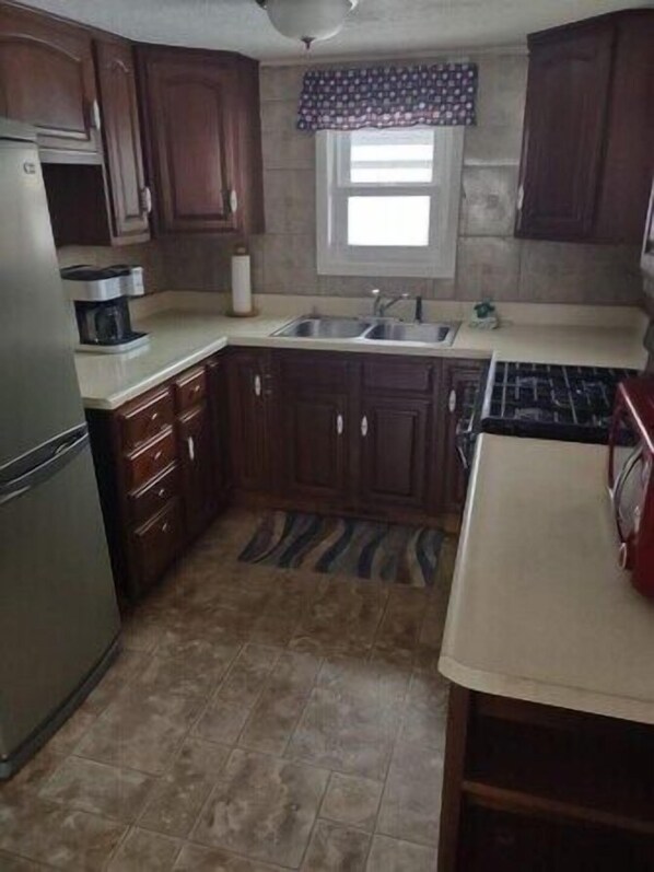 A compact kitchen with dark wooden cabinets, a stainless steel refrigerator, a coffee maker, a sink, and a patterned window valance. - Cozy two bedroom bungalow (Duluth)