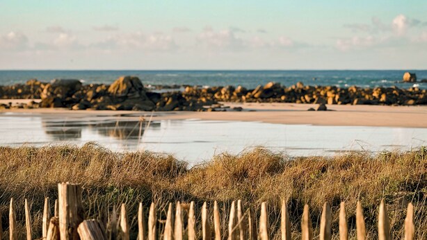 A beach with a wooden fence in the foreground, a rocky shoreline, and calm water. - Exceptional villa with indoor pool. beach 300m away, sleeps 14. (Cléder)