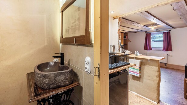 A rustic bathroom with a stone sink, wooden shelves, and a wooden door leading to a kitchen area with a window and curtains.