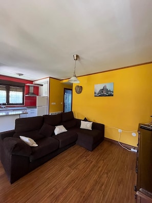A living room with a sectional sofa, a ceiling fan, a clock on the wall, and a kitchen in the background.