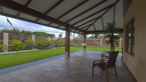 Eine überdachte Terrasse mit zwei Hängematten, einem Stuhl und Blick auf eine grüne Wiese und Bäume.