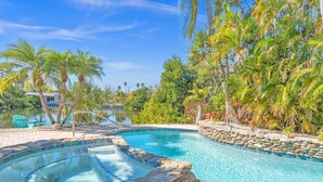 Piscina com hidromassagem, cercada por palmeiras e vista para uma casa do outro lado da água.