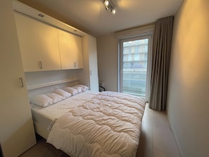 A bedroom with a large bed, built-in wardrobe, and a window with curtains. - Apartment in Nieuwpoort With Balcony (Nieuwpoort)