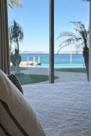 A bedroom with a large window offering a view of a beach and ocean.