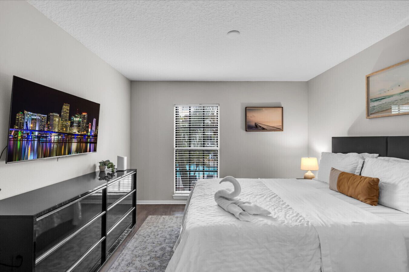 A hotel room with a bed, a desk, a TV, a window with blinds, and a painting on the wall.