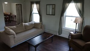 A living room with a beige sofa, a dark wooden coffee table, and a brown armchair. There is a window with curtains, a lamp on a side table, and a dining area in the background.