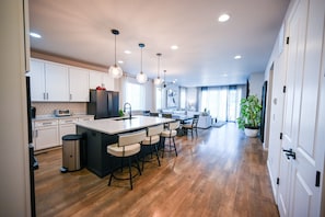 A modern kitchen with a central island, bar stools, pendant lighting, and a view to the living area. - Lux Family Retreat|Pool Table|13 Min to Airport (Commerce City)