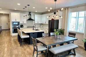 A modern kitchen with white cabinets, a central island, a dining table with benches, and a large window with blinds. - Lux Family Retreat|Pool Table|13 Min to Airport (Commerce City)