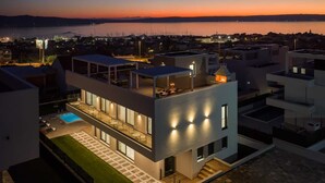 Un edificio moderno de varios pisos con piscina en la azotea y zona de estar al aire libre, con vistas a una masa de agua al atardecer.