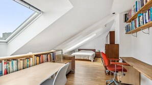 A modern loft bedroom with a sloped ceiling, a wooden desk, a red office chair, a bed, and a bookshelf filled with books.
