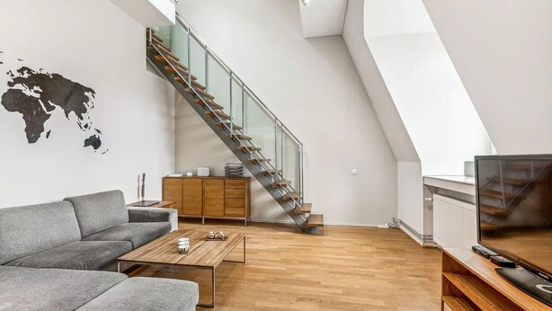 A modern living room with a grey sofa, wooden coffee table, and a TV on a wooden stand. A staircase with glass railings leads to an upper level.