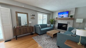A living room with a sofa, coffee table, and fireplace. There is a television mounted above the fireplace. - Serene Lake Ontario Retreat • Entire Home Near Trails & Beach. (Ajax)