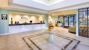 A hotel reception area with a reception desk, a glass-top table with a vase of flowers, and a seating area with a sofa and armchairs.