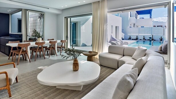 A modern living room with a dining area, a large sofa, and a view of a pool and building. - Walk to Naousa | 5BR Pool Villa | Villa Yagha (46CJ+P7Q Naousa, Greece)