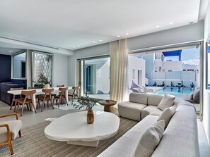 En moderne stue med spiseområde, stor sofa og udsigt til pool og bygning.