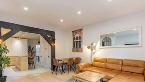 A modern living room with a sofa, dining table, chairs, and a kitchen area visible through an open archway.