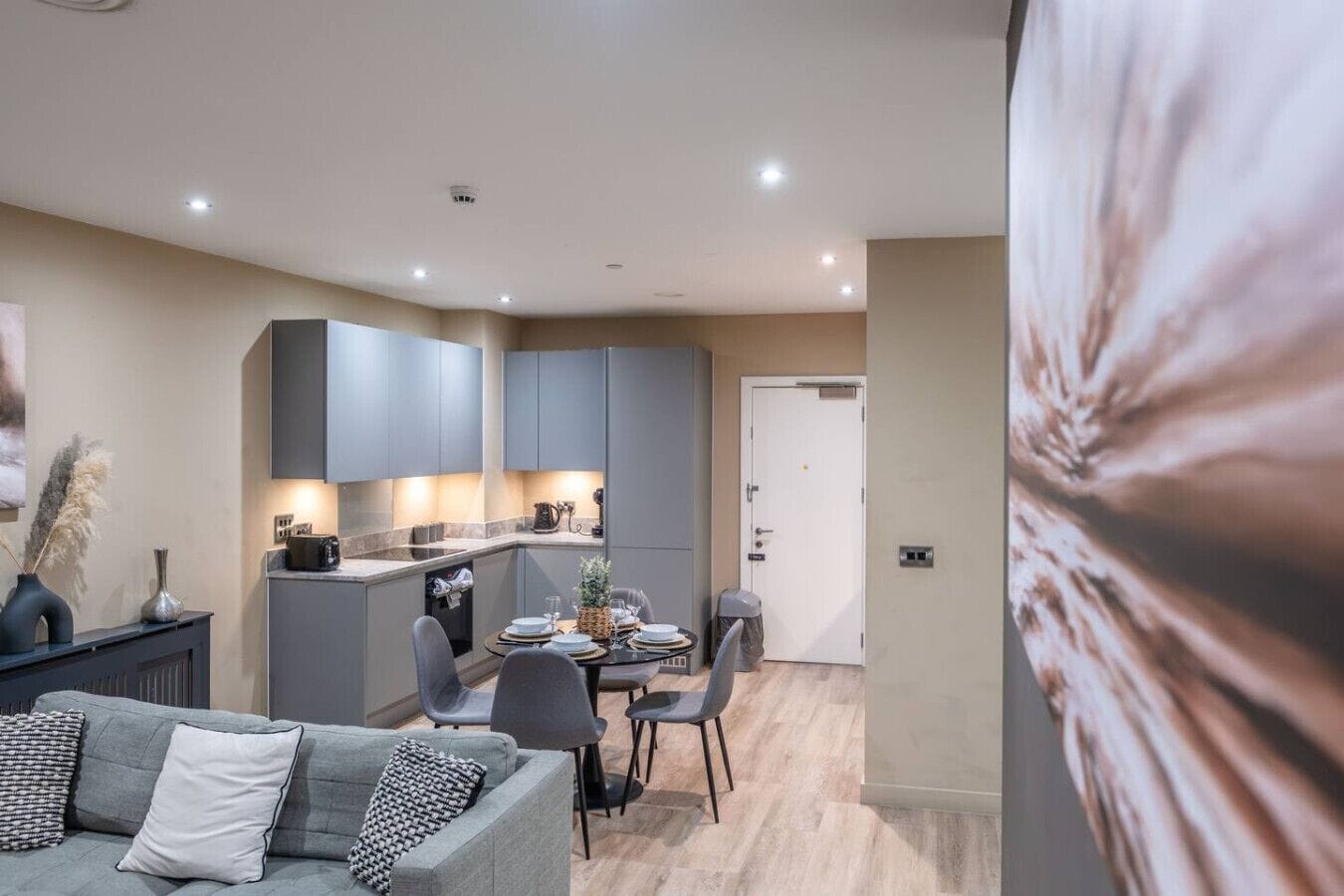 A modern kitchen with grey cabinets, a dining area with a table set for four, and a grey sofa with patterned cushions.