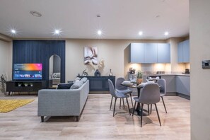 A modern living room with a grey sofa, dining table, and chairs, a flat-screen TV, and a kitchen area with cabinets and a countertop. - Central Manchester Townhouse + Free Parking. (Manchester)