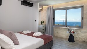 A modern bedroom with a large bed, a bedside table with a plant, and a view of the sea through a large window. - White Stone Elegance with Pool, Jacuzzi & Golden Sunsets (Chania)