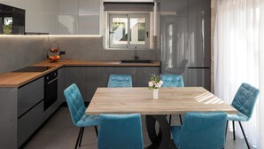 A modern kitchen with a wooden countertop, a built-in oven, and a sink. There is a dining table with four blue chairs and a vase with flowers. - White Stone Elegance with Pool, Jacuzzi & Golden Sunsets (Chania)