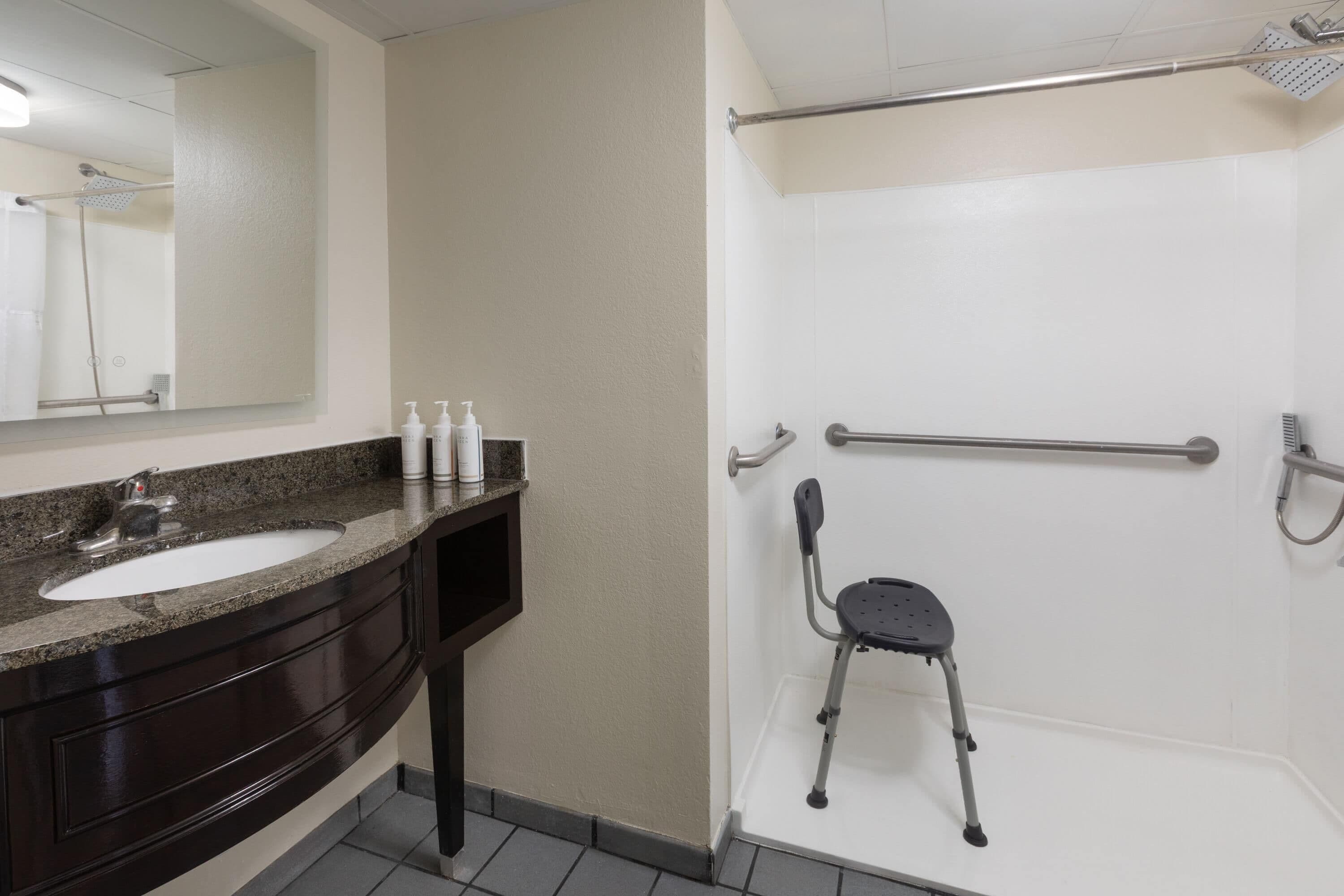 A bathroom with a sink, mirror, toiletries, a shower with a grab bar, and a wheelchair-accessible seat.