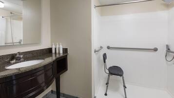 A bathroom with a sink, mirror, toiletries, a shower with a grab bar, and a wheelchair-accessible seat.