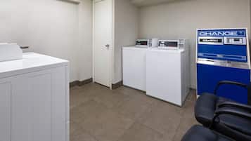 A laundry room with two washing machines, a change machine, and a chair.