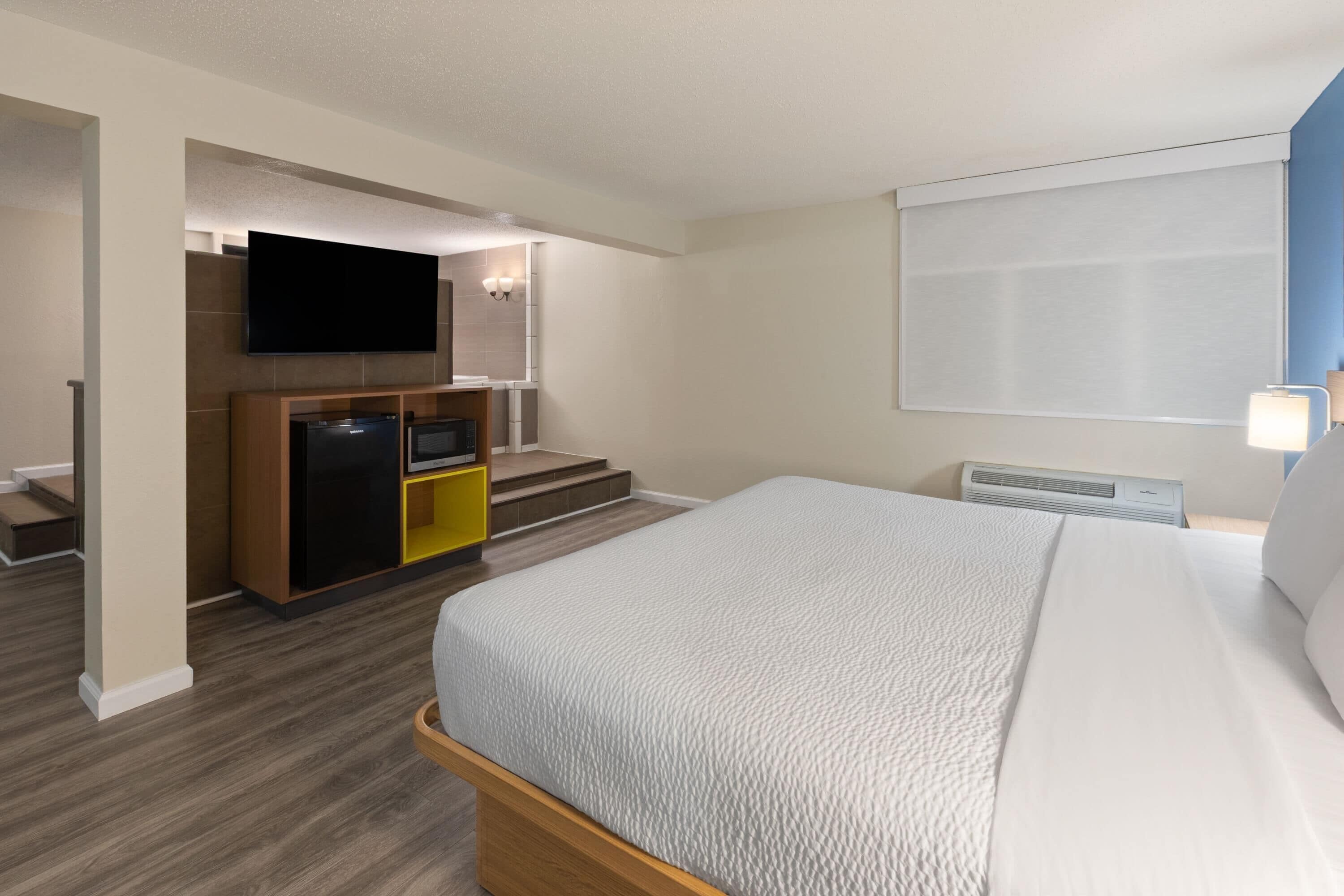 Une chambre d’hôtel avec un lit, un téléviseur fixé au mur, une petite kitchenette et une fenêtre avec des stores.
