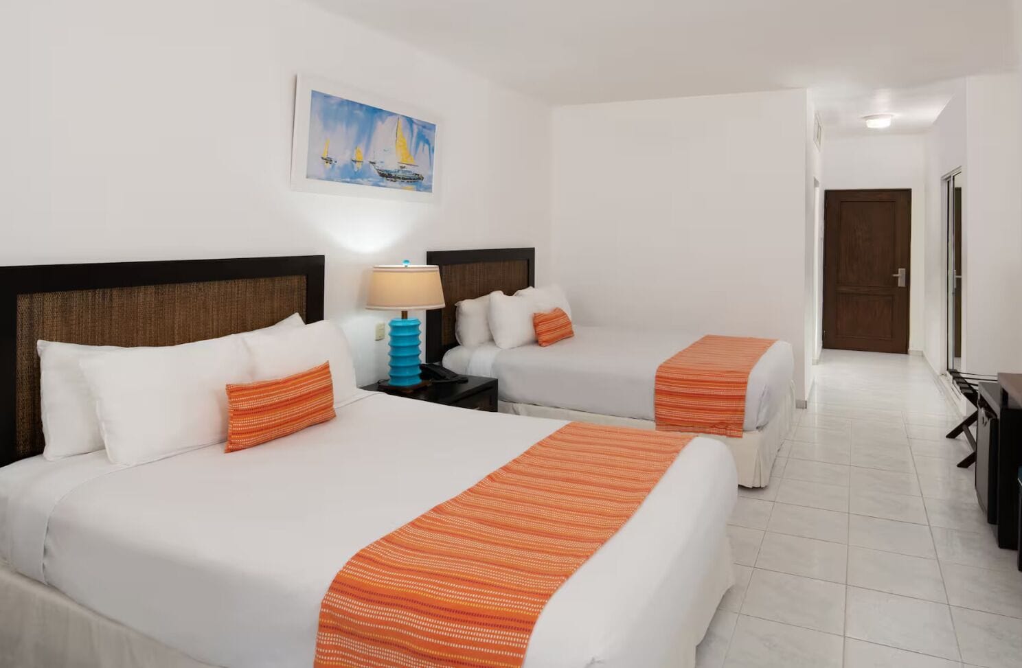 A hotel room with two beds, a painting of sailboats, a lamp, and a door leading to another room.