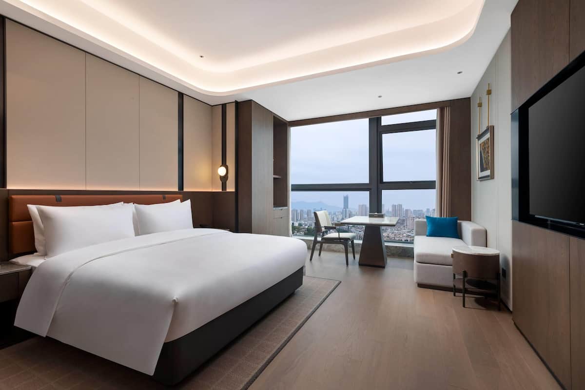 Una habitación de hotel moderna con una cama grande, un área de descanso con una mesa, un televisor y vistas a la ciudad.
