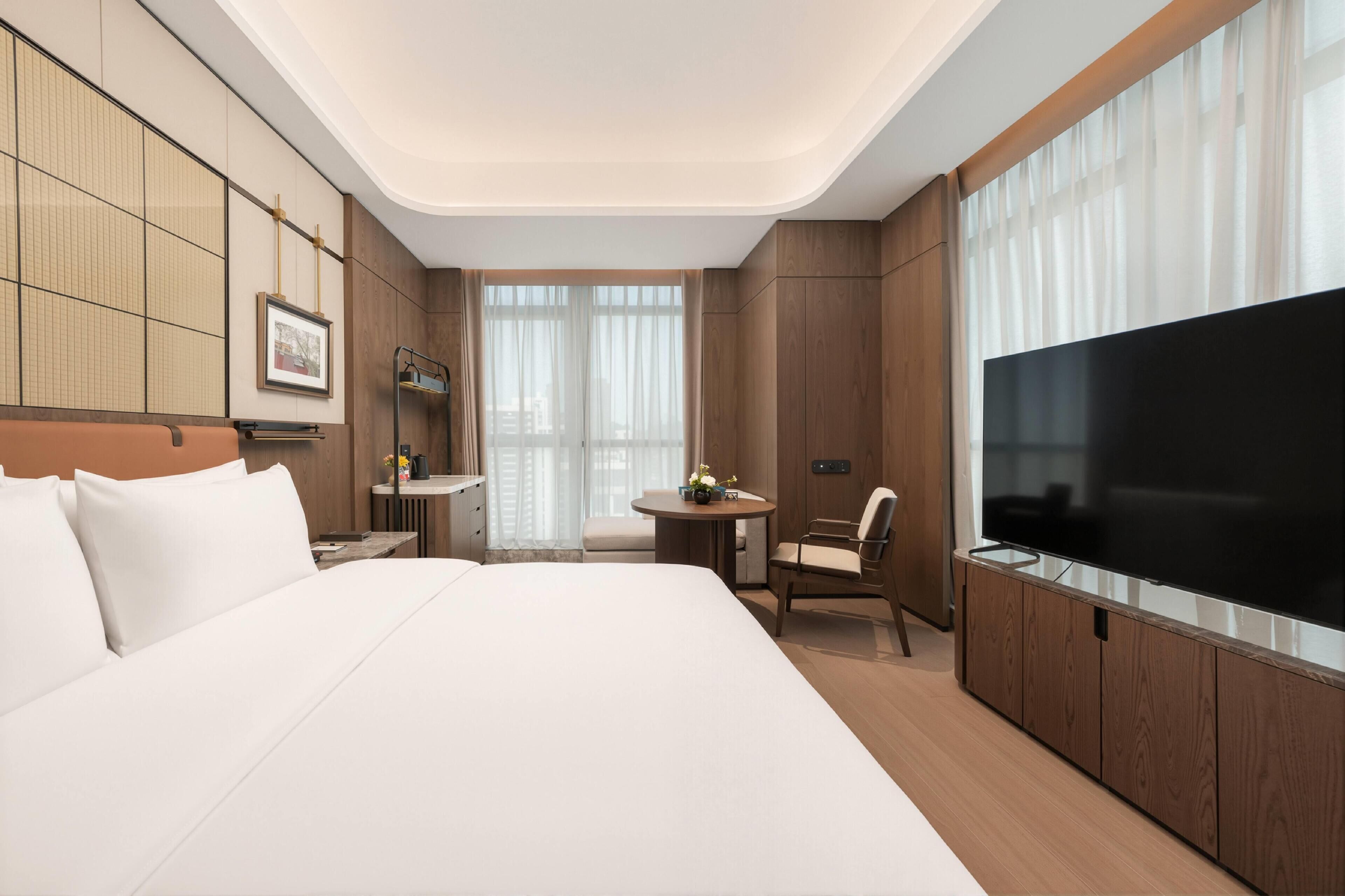 A hotel room with a large bed, a TV, a small table with a chair, and a window with curtains.