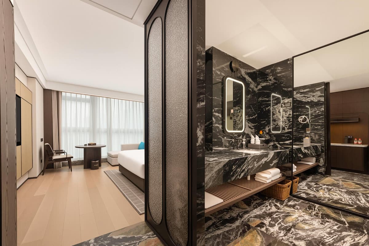 Una habitación de hotel moderna con una cama grande, un área de descanso con una mesa y sillas, un baño con tocador y espejo de mármol, y un ventanal con cortinas.