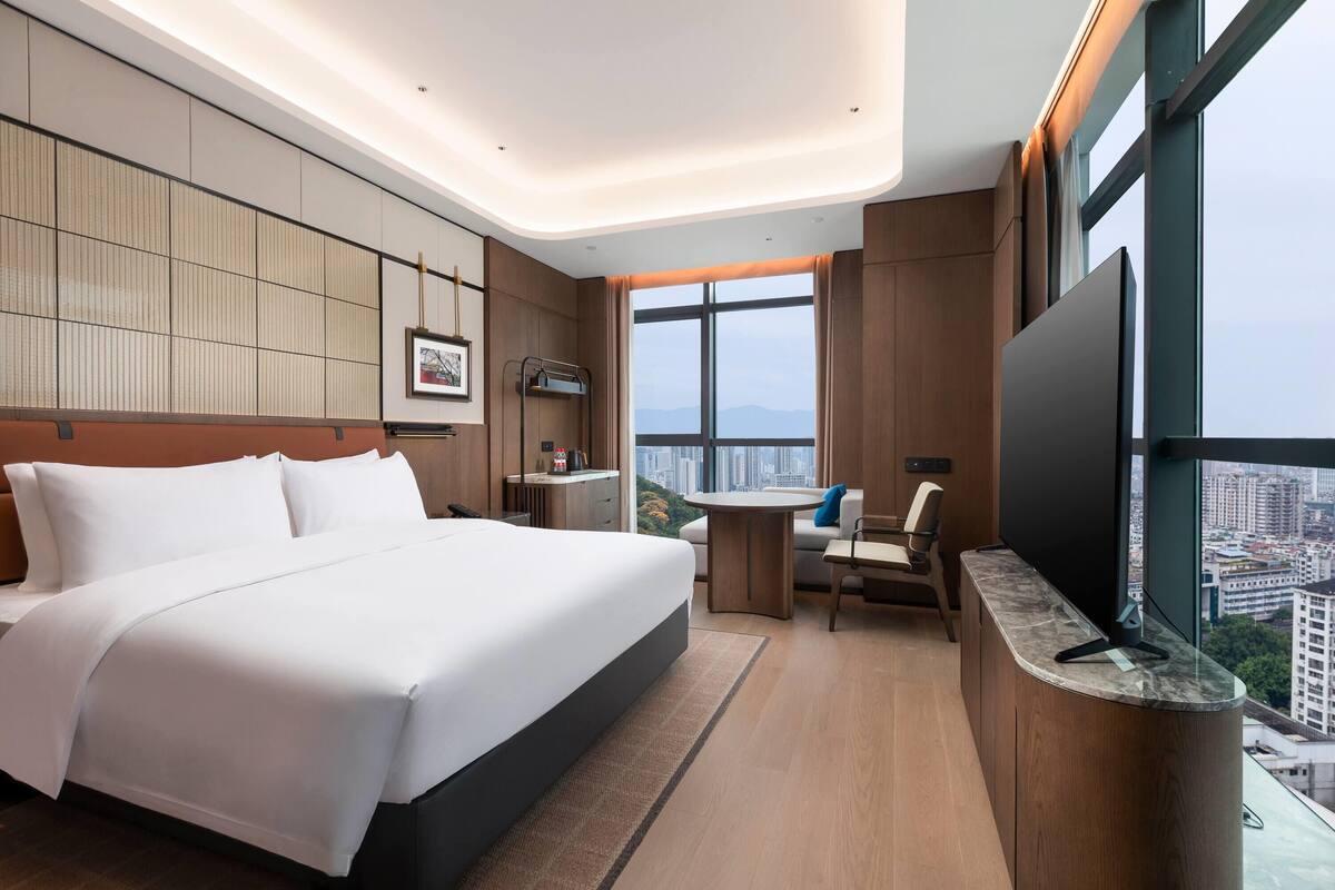 Una habitación de hotel moderna con una cama grande, un escritorio, una silla, un televisor y vistas a la ciudad.