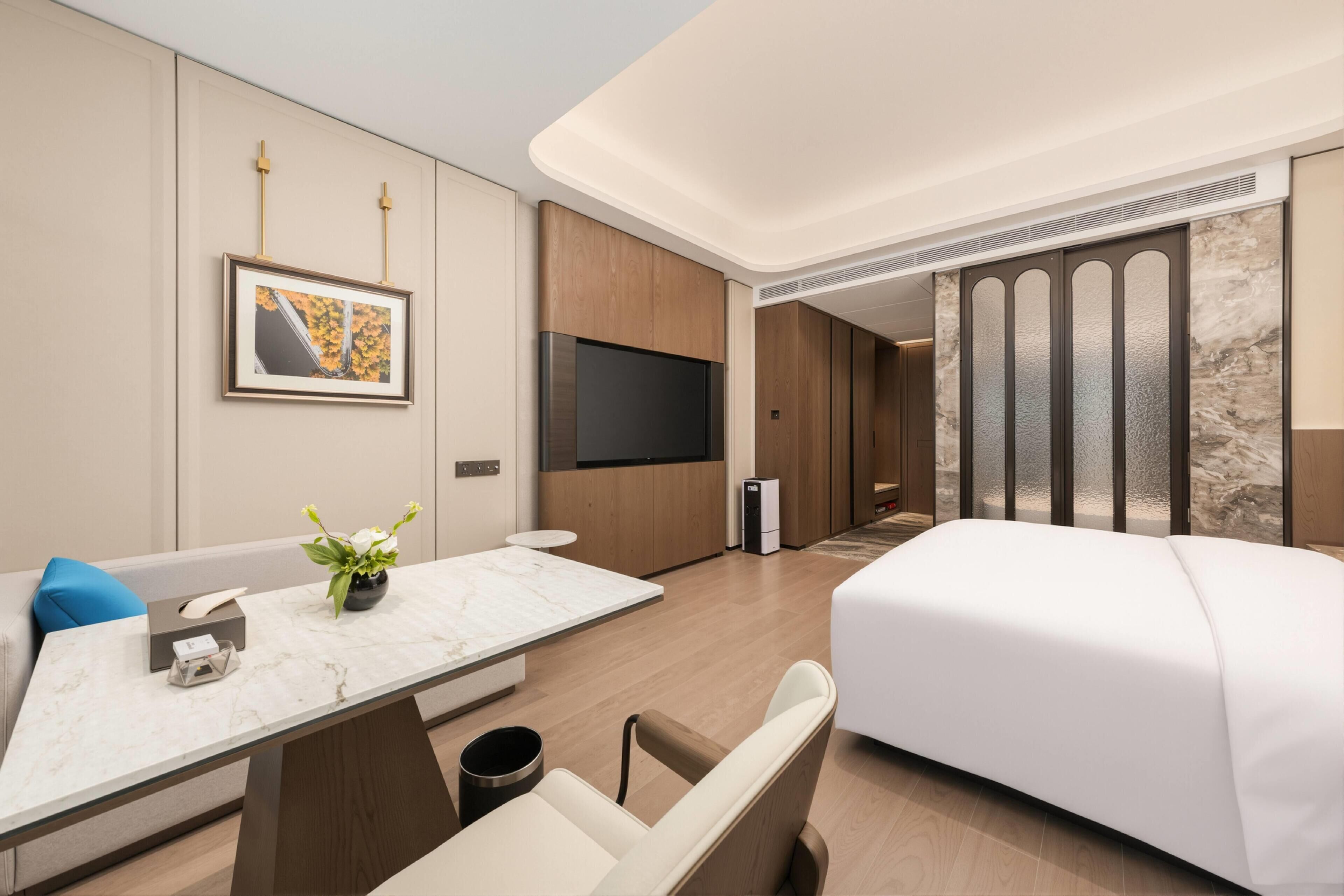 Una habitación de hotel moderna con cama, televisión, escritorio y un florero, y un ventanal amplio.