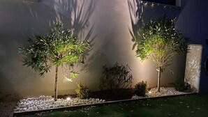 Twee verlichte bomen in een tuin met witte kiezelstenen en groen gras, voor een witte muur met een raam.