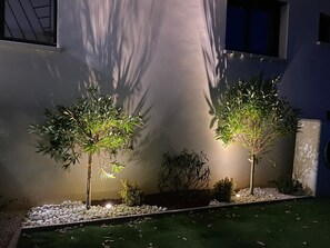 Zwei beleuchtete Bäume in einem Garten mit weißen Kieselsteinen und grünem Gras, vor einer weißen Wand mit einem Fenster.