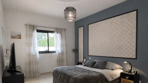 Een moderne slaapkamer met een groot bed, een televisie op een standaard, een nachtkastje met een lamp en een groot raam met gordijnen.