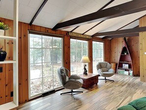 A cozy cabin interior with wooden walls, large windows, two armchairs, a small table, and a bookshelf. - Blue Haven Retreat (Oakland)