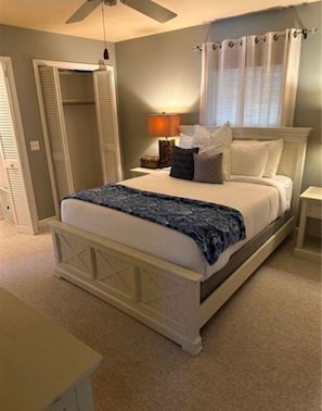 A bedroom with a bed, a nightstand, a lamp, a window with curtains, and a ceiling fan. - The Blue Retreat in Fort Myers Beach (Fort Myers Beach)